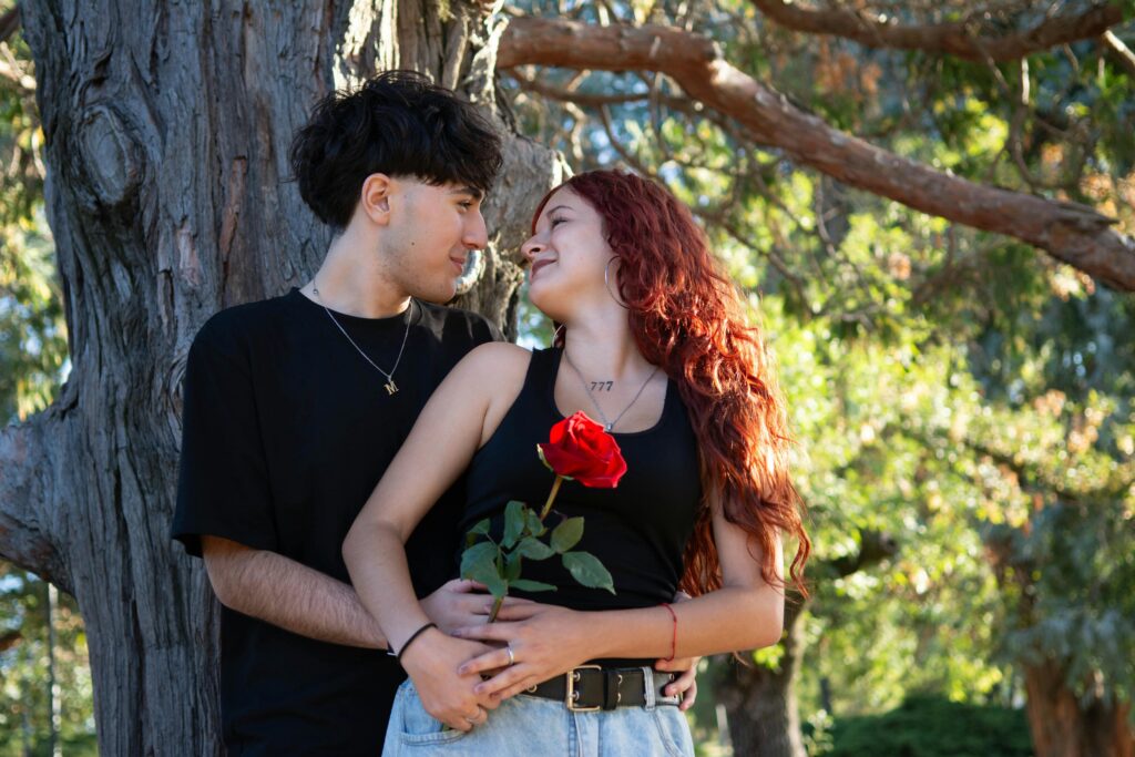service-1 A young couple embraces lovingly under a tree, holding a red rose in a serene outdoor setting.