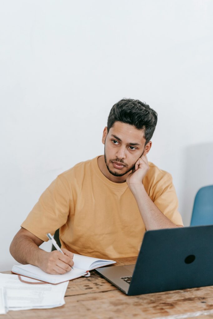 Job Problem Solution Pensive young Hispanic male entrepreneur in casual clothes thinking about business strategy while sitting at table with laptop and writing in notebook